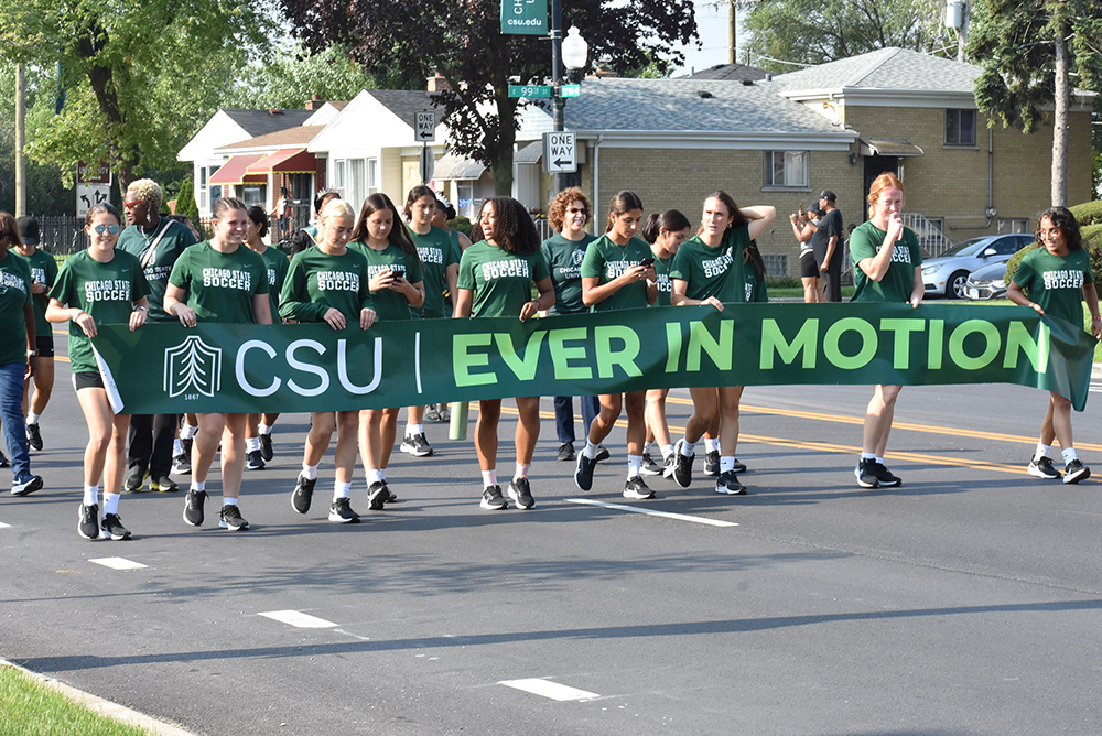 Approximately 20 members of  #the Chicago State University soccer team walk in the street during the annual Back to School parade. They are holding a long, dark green banner with the university logo and the letters CSU in white, and the words “Ever in Motion” in bright green.