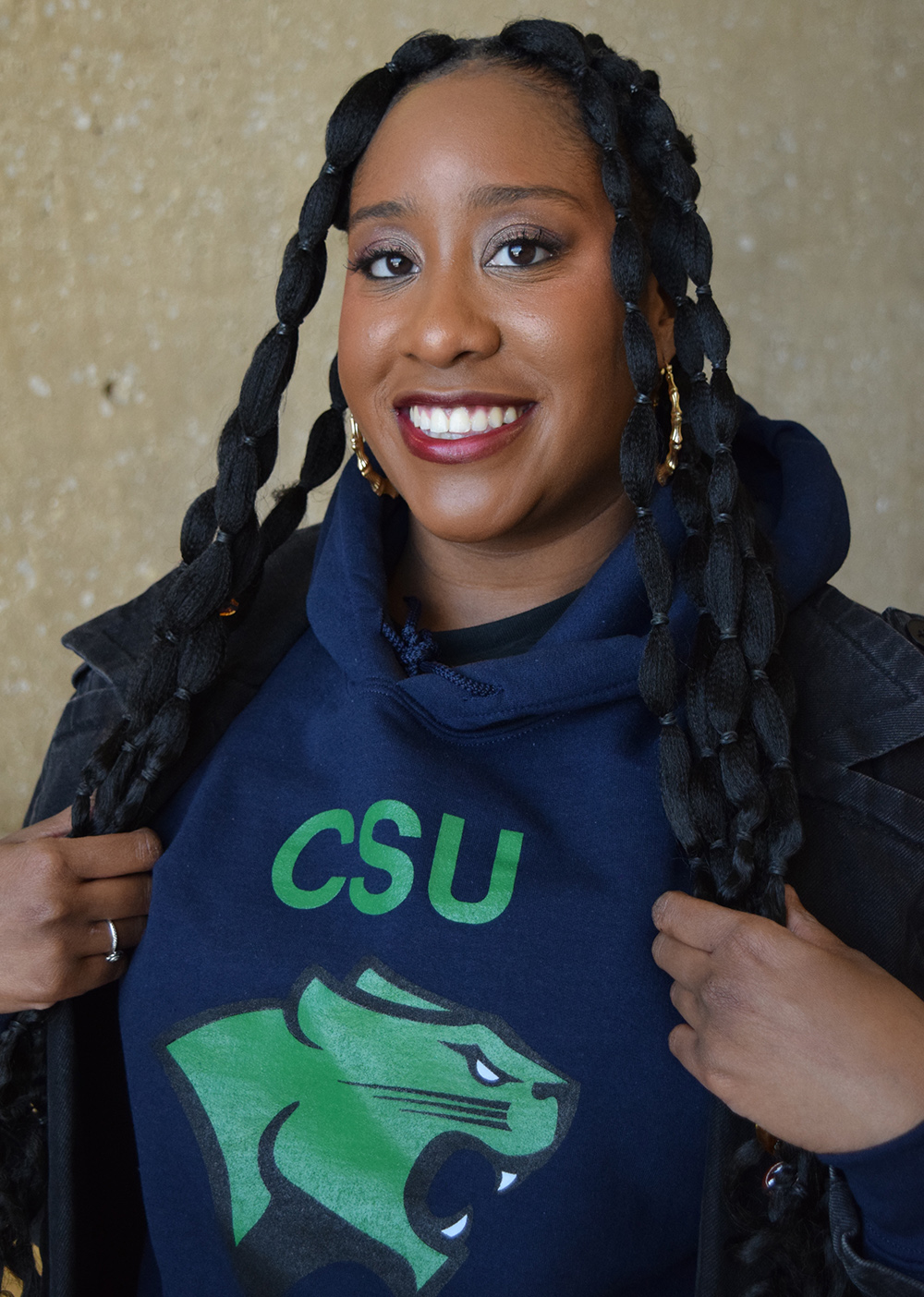 A young woman with long, rubber-banned hair, smiles into the camera. She’s wearing a dark, blue hoodie with CSU written in large green letters across the front, and an image of the Cougar mascot below it.
