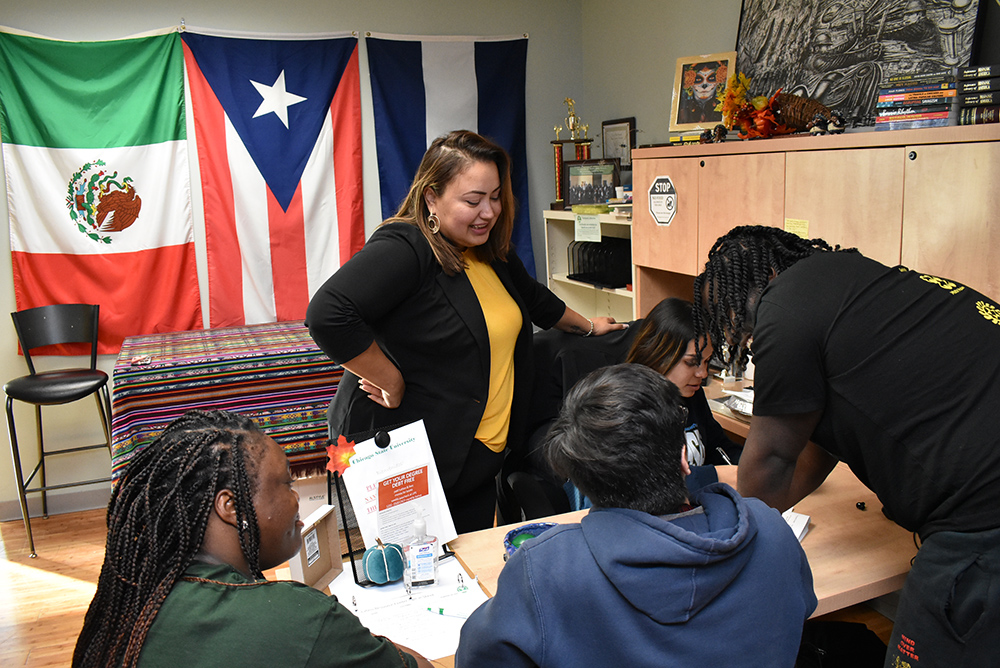A group of students in the Center for Access, Support & Advancement (CASA) office work collaboratively around a small table while the director provides support. In the background, the wall is decorated with the Mexican and Puerto Rican flags.