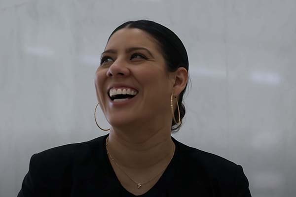 A close-up, candid portrait of a woman with dark hair pulled back, wearing large gold hoop earrings and a black blazer, laughing joyfully while looking toward the upper left.