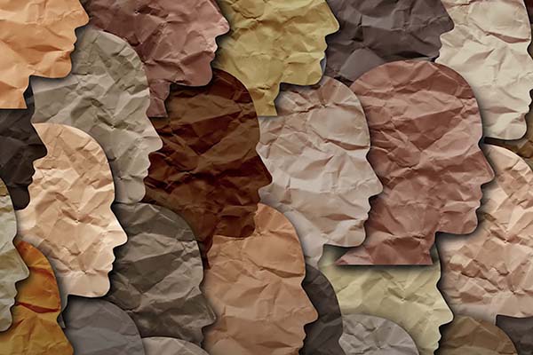 A stylized graphic illustration composed of numerous profile silhouettes of human heads in various skin tones. Each silhouette features a crumpled, textured paper effect, overlapping to create a mosaic pattern.