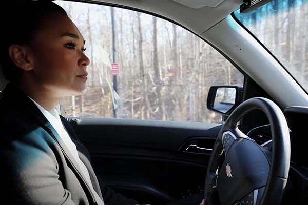 A side-profile view of a woman with her hair in a bun sitting in the driver’s seat of a Chevrolet vehicle. She is looking out the windshield at a wooded area through the driver's side window.