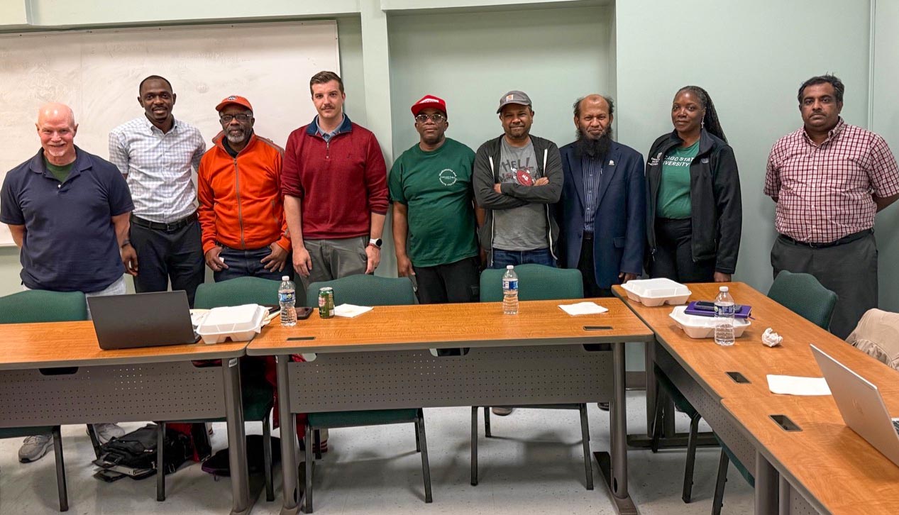 Nine people, consisting of Chicago State University alumni, staff and faculty members standing side by side behind brown tables and green chairs.