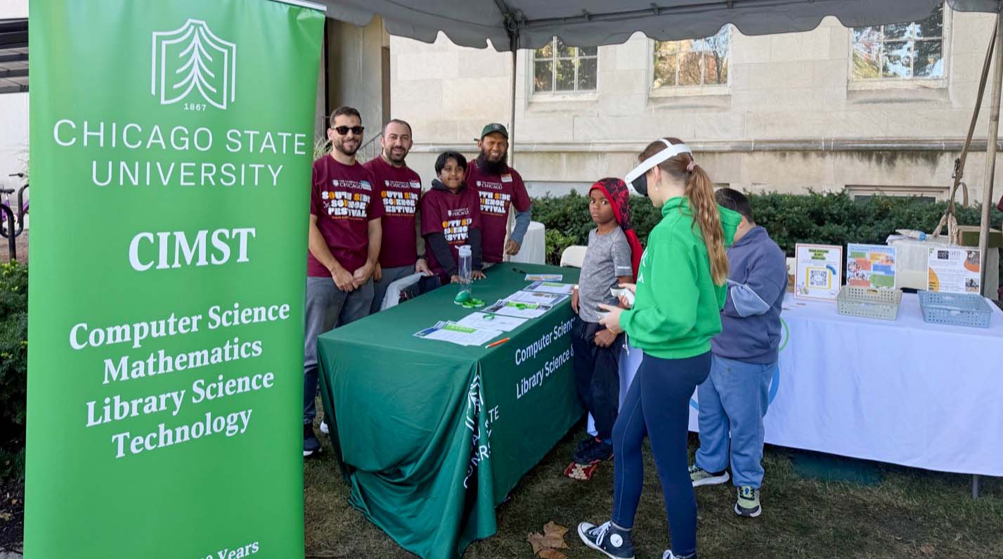 In the foreground on the left stands a tall, green CSU banner for CIMST. In the background, students from CIMST, the CIMST department chair and children pose behind a table with swag items.