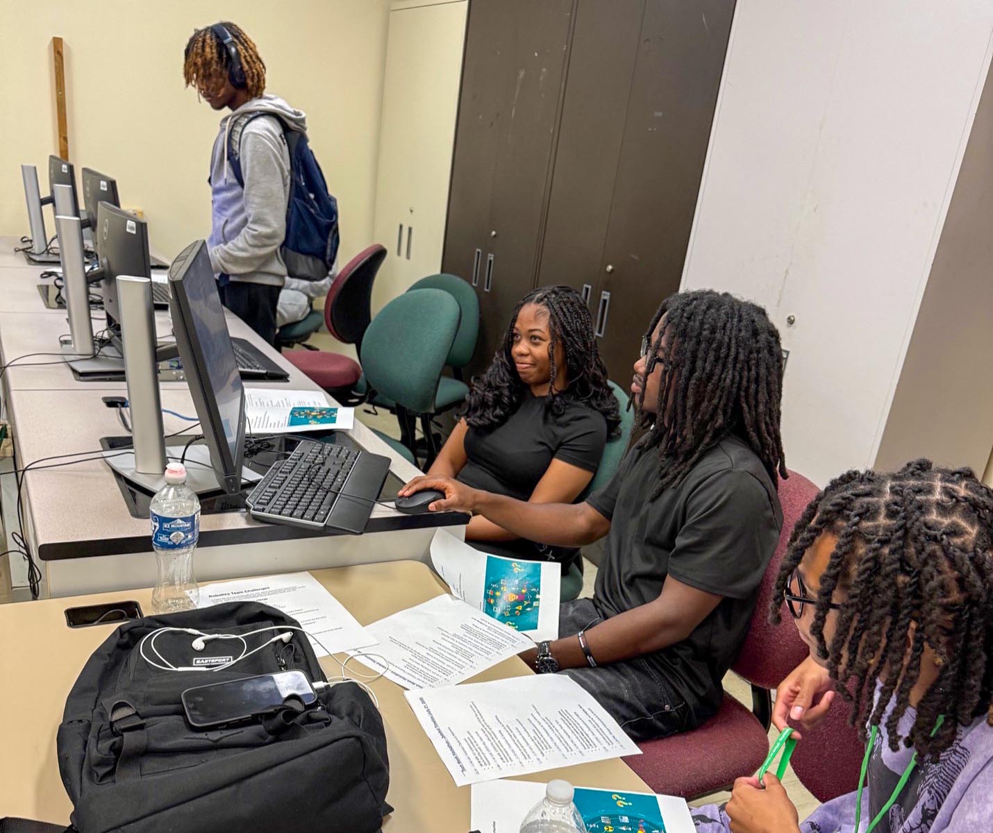 A young man with locs uses a computer mouse while looking at a monitor alongside a young woman in a computer lab. Printed handouts, a black backpack, a smartphone, and a water bottle sit on the desk in front of them.