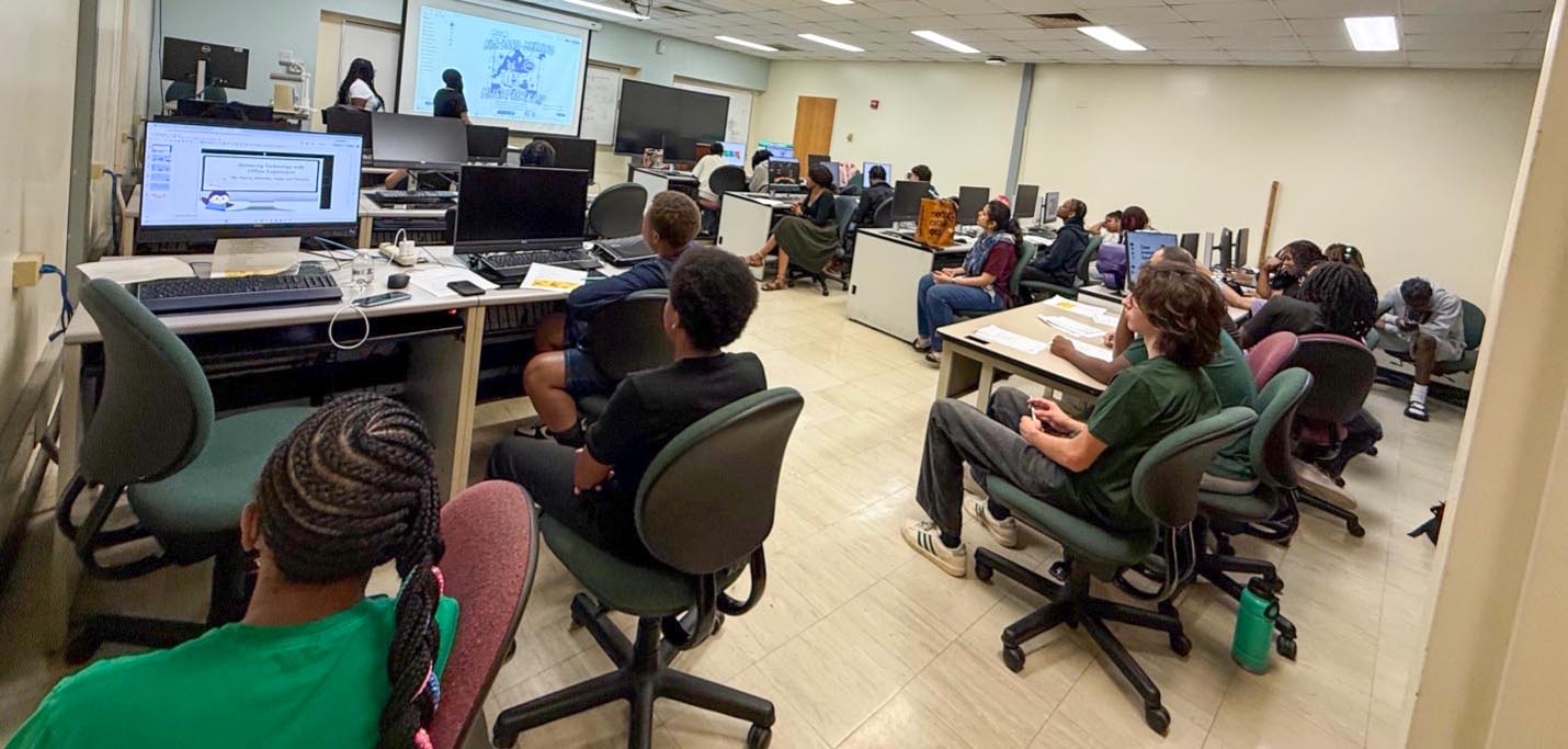 Students seated at desks in a computer lab view a slide presentation displayed on a large screen at the front of the room.