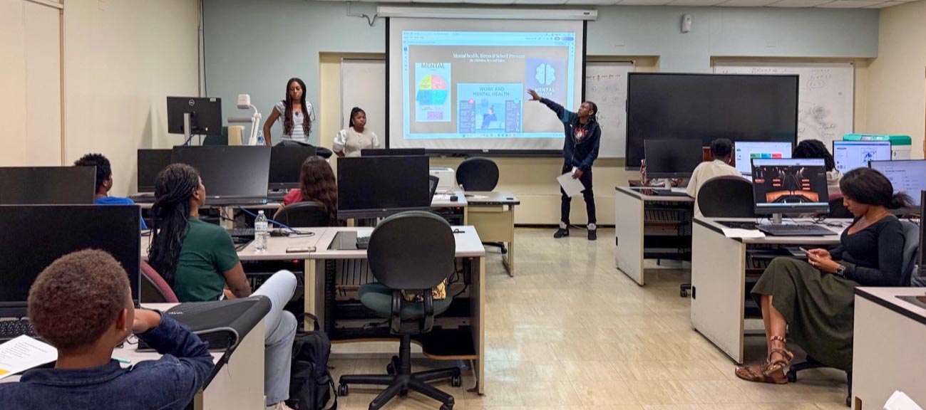A student points to a slide presentation on mental health while co-presenting in front of a computer lab classroom.