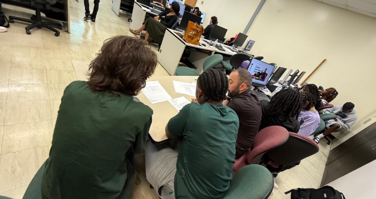 View from behind a group of students seated around a shared table alongside an instructor reviewing papers, with additional computer workstations and students visible in the background.