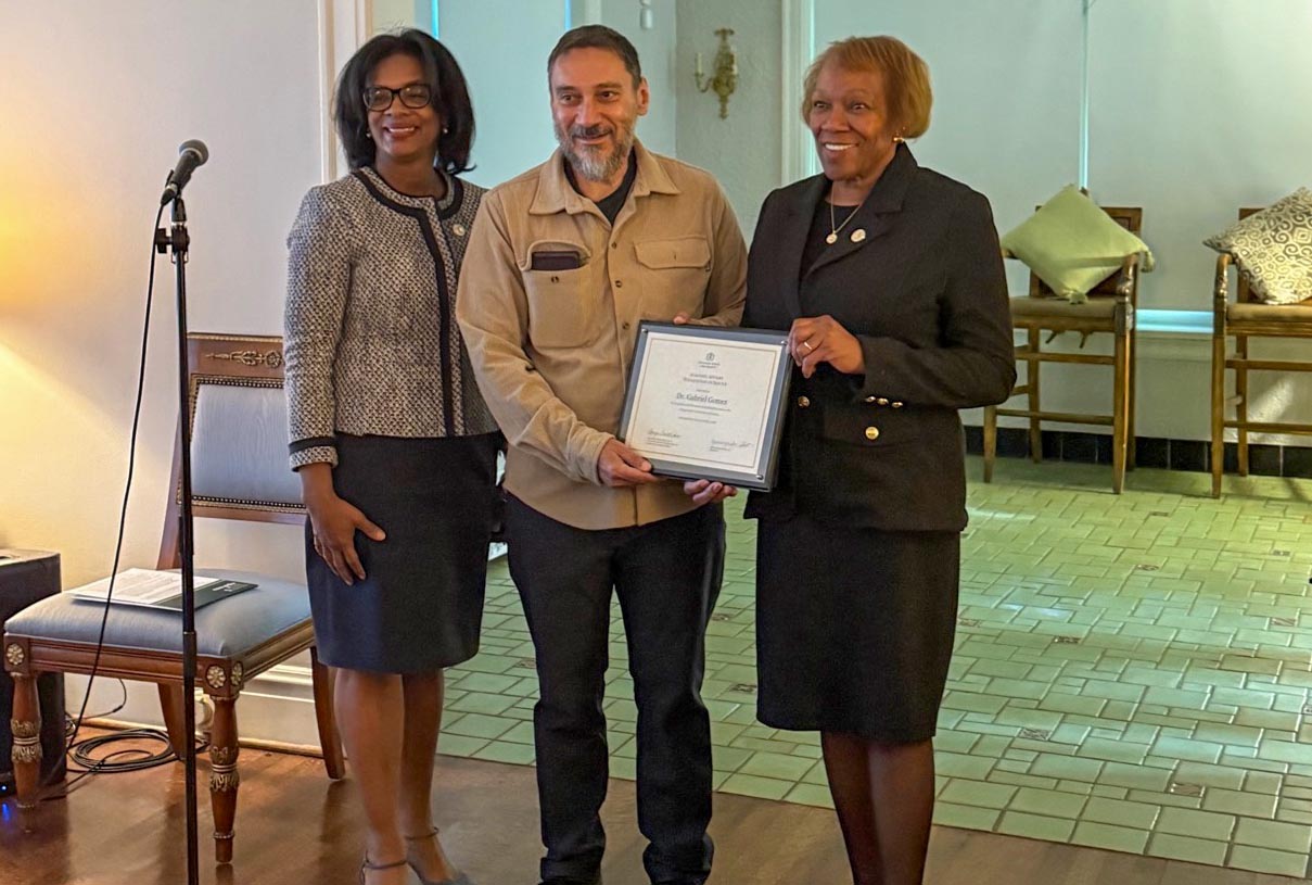 Dr. Gomez accepts a certificate of appreciation from Provost Sonja Feist-Price and President Scott during a retirement celebration. On the left is a microphone and an ornate antique chair. In the background, a tiled floor is visible.