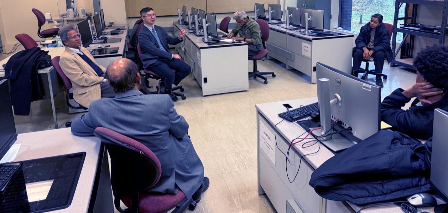 A group of people sit in a computer lab, listening while a man in dark blue suit speaks.