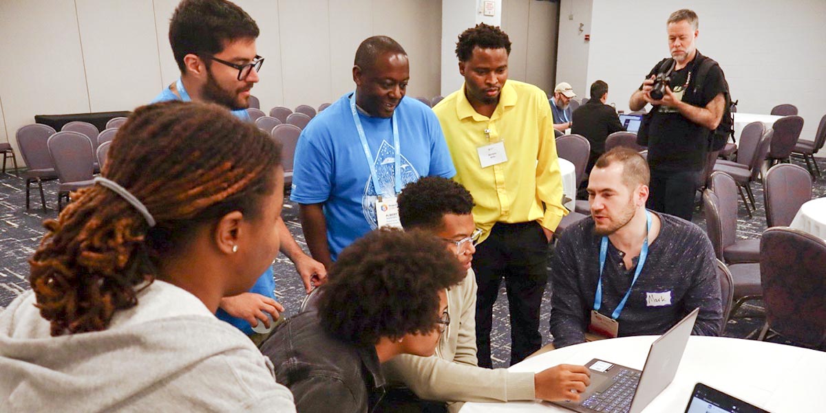 Seven conference attendees gathered behind a laptop, working together.