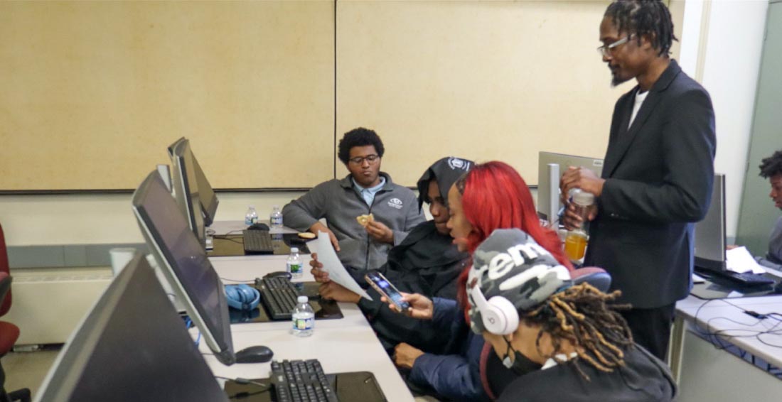 A CIMST student stands behind four high school students sitting in front of monitors in a computer lab.