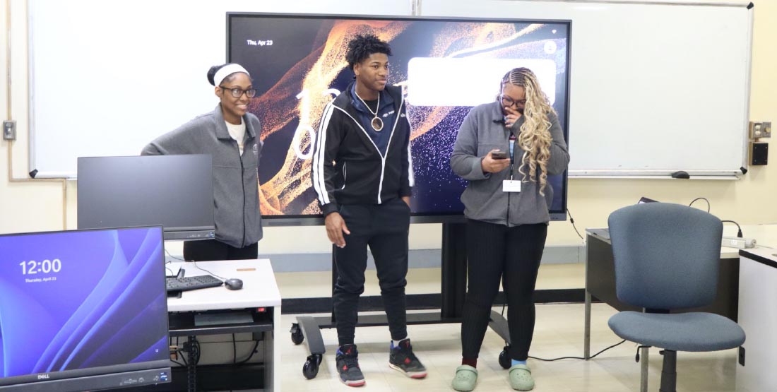 Three high school students stand in front of a smart board smiling.