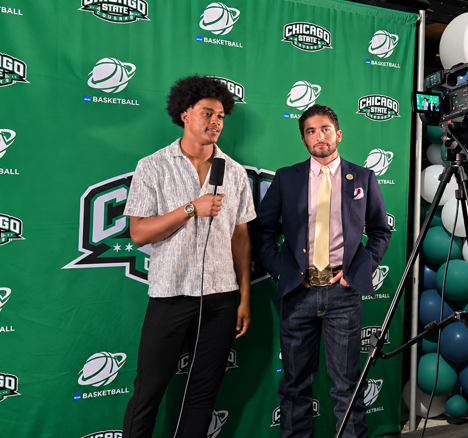 A student interviewer holding a microphone speaks with a man in a blazer standing in front of a green Chicago State athletics step-and-repeat backdrop.