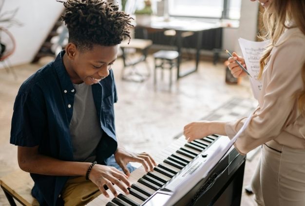 A young person is smiling while playing a keyboard with the help of an instructor standing over them.