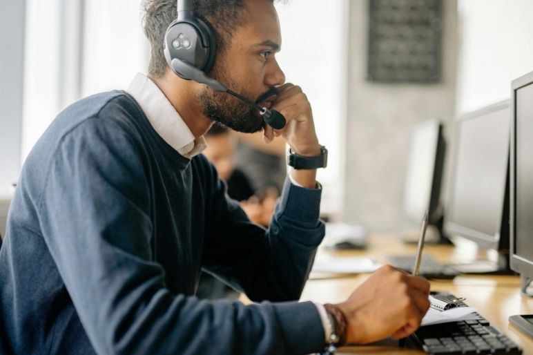 A man with a beard and headset is sitting at a computer, holding a pen and looking intently at the screen.
