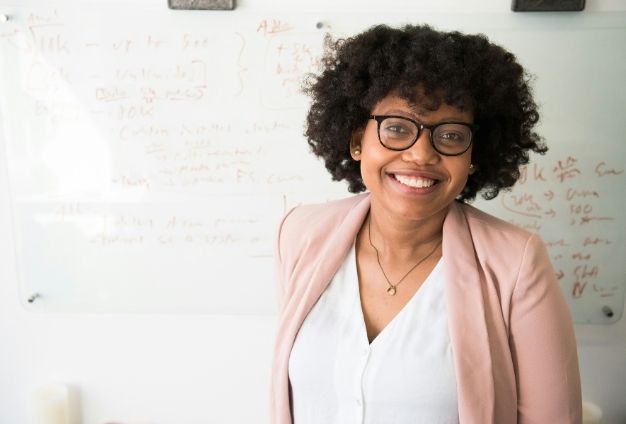 A woman with glasses and a pink blazer is smiling at the camera, standing in front of a whiteboard with handwritten notes