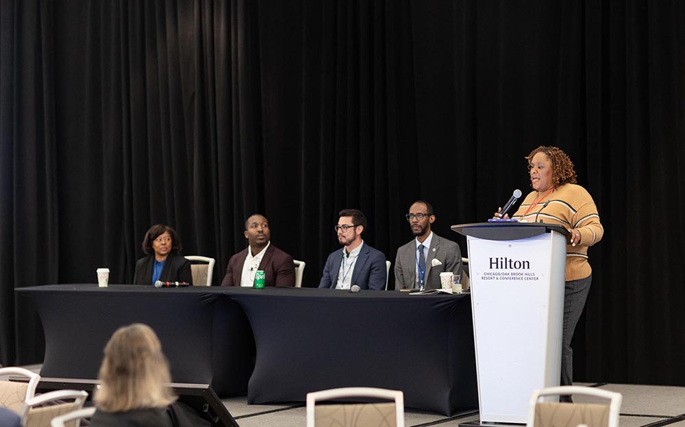 A panel discussion in a conference room with a black curtain backdrop. Four panelists (two women, two men) are seated at a table covered in a black tablecloth. On the far right, a woman with curly hair is speaking at a white podium.