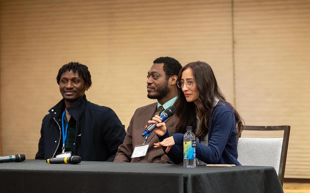 Three student researchers sit behind a table with a black tablecloth during a panel discussion. One of the students, with long, dark hair and glasses is speaking into a microphone.