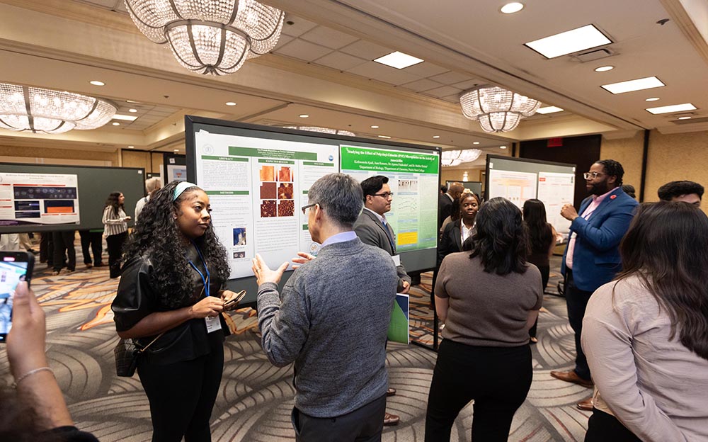 A wide shot of people in a conference hall where multiple research posters are being presented. In the foreground, two people are talking while others present their research in the background.