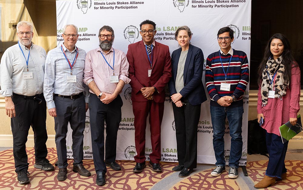 Seven people stand next to each other, posing for a photo in front of a white backdrop with the ILSAMP logo printed across.
