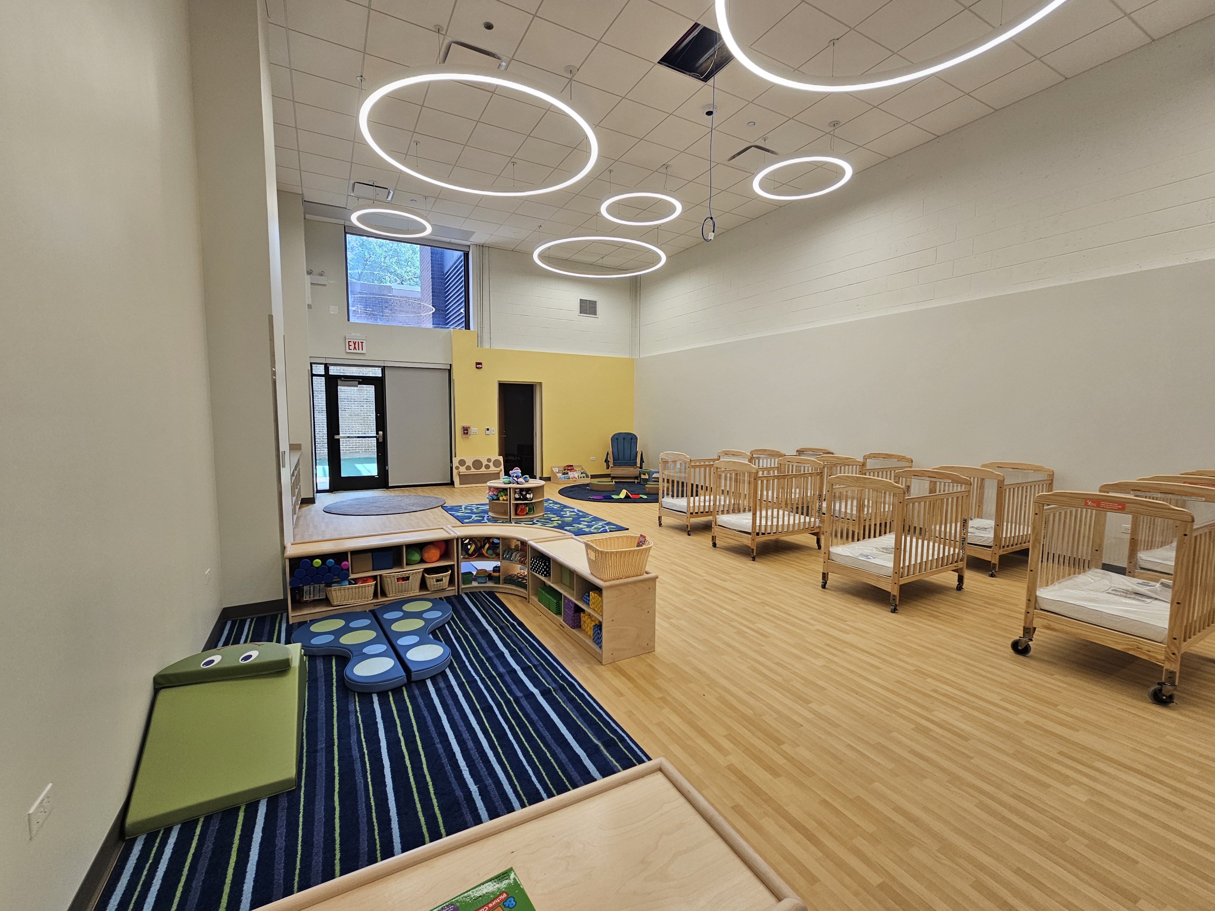 A spacious infant room complete with light, wood-colored padded floors and soft area rugs. A play area with a blue rug includes low shelving with storage bins of infant toys. Several small wooden cribs with mattresses are in the center of the room and a rocking chair sits in the corner.