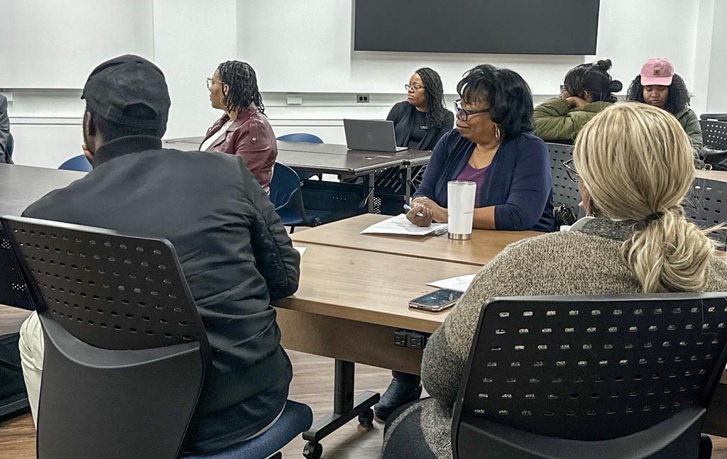 Attendees sitting at a table facing a speaker during an industry day event, with one woman taking notes