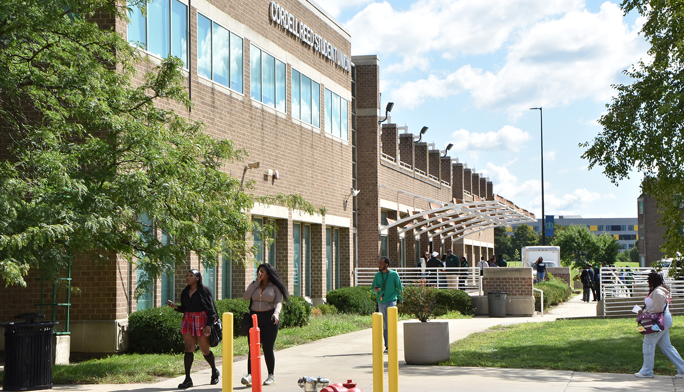 A sun-filled wide-angle photo of the Cordell Reed Student Union building on the left with a blue sky and bright, white clouds at the top right, and trees on both sides. Several students can be seen walking in multiple directions in the foreground while a group of students are socializing in the background.