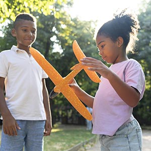 A sunny photo of a young boy and girl standing outside. The girl is holding an orange model airplane. 